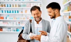 A friendly male pharmacist in a white coat explains a medication to a smiling, middle-aged male customer. The pharmacist holds a small white box and points to it as the customer looks on attentively.