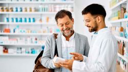 A friendly male pharmacist in a white coat explains a medication to a smiling, middle-aged male customer. The pharmacist holds a small white box and points to it as the customer looks on attentively.