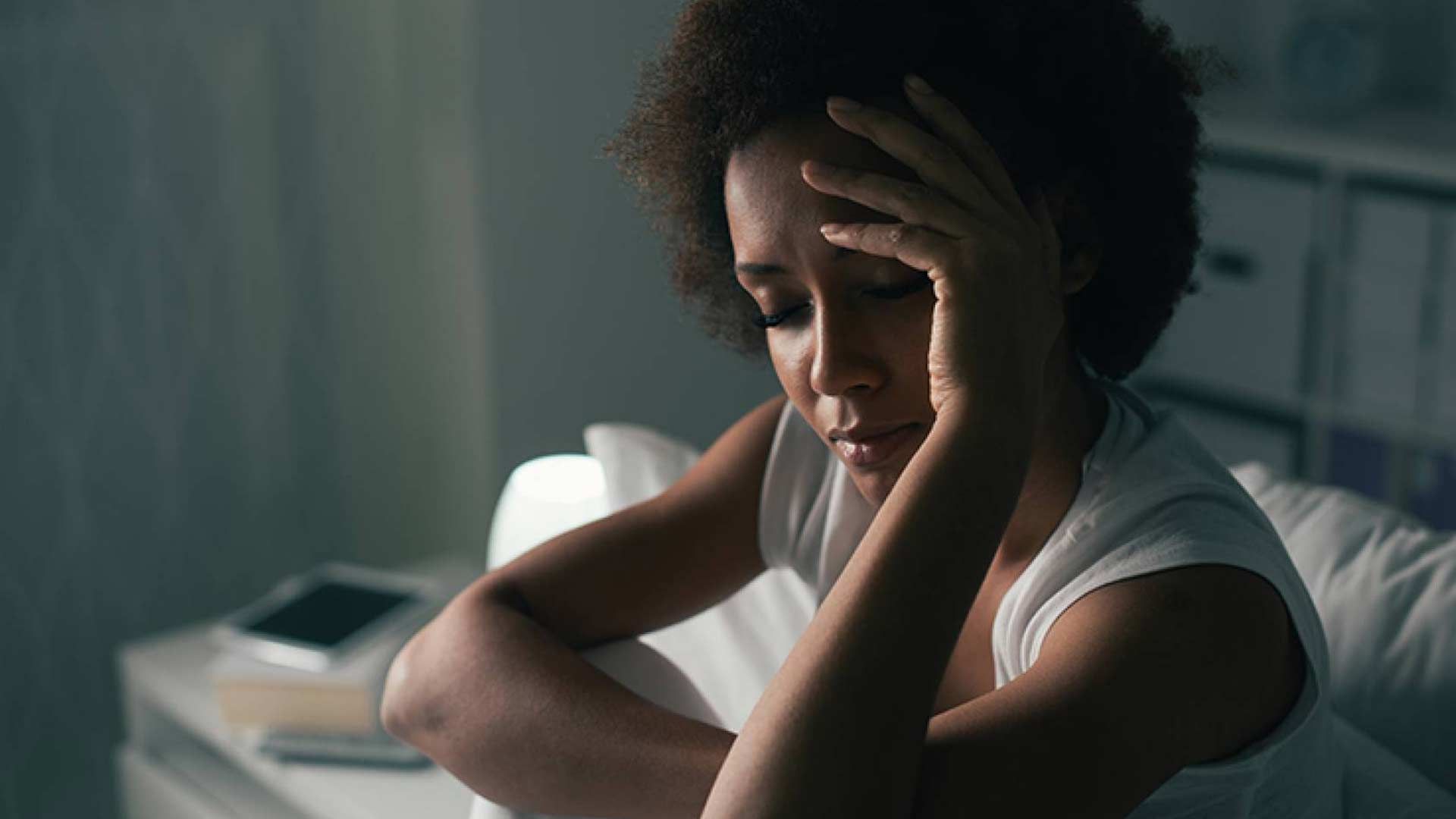 A Black woman sits on the edge of her bed in a dimly lit room, appearing distressed. With a sad expression, she rests her head in one hand, looking downwards as if deep in thought or struggling with insomnia.