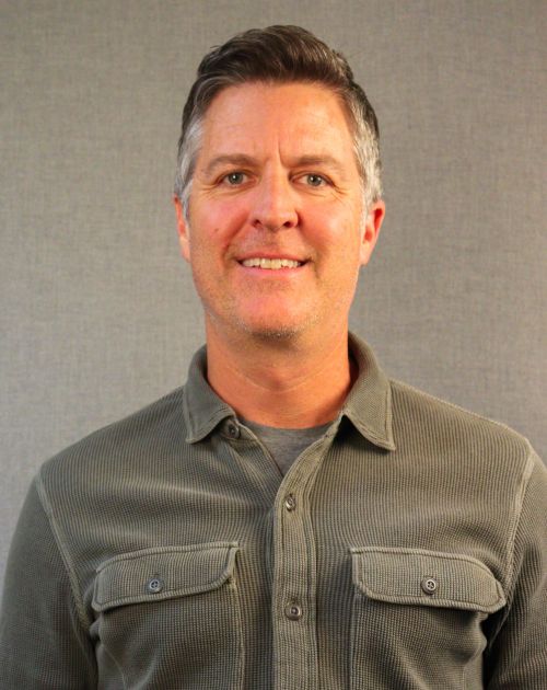 A headshot of Eric Roberts, a man with short dark hair and glasses. He is smiling warmly at the camera
