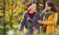 A caring younger woman in a yellow coat walks arm-in-arm with a smiling elderly woman through a park in the autumn.