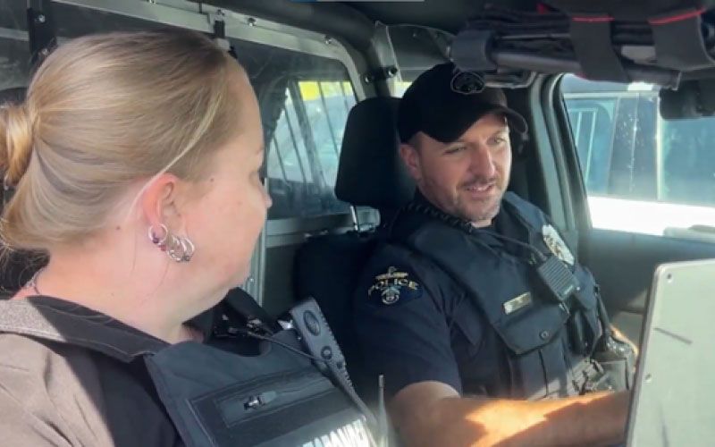 A male police officer and a female co-responder are sitting together in the front seat of a patrol vehicle.