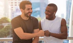 Two diverse friends are laughing warmly while giving each other a fist bump in a bright, urban outdoor setting. One man wears a black t-shirt, while the other man, who is Black, wears a white sleeveless athletic shirt and has earbuds in.