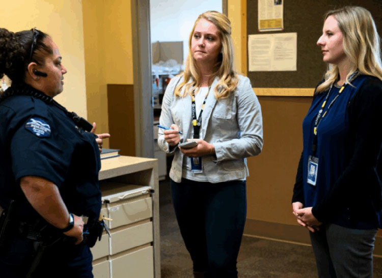 A female police officer in uniform stands and speaks with two female co-responders in an office setting.