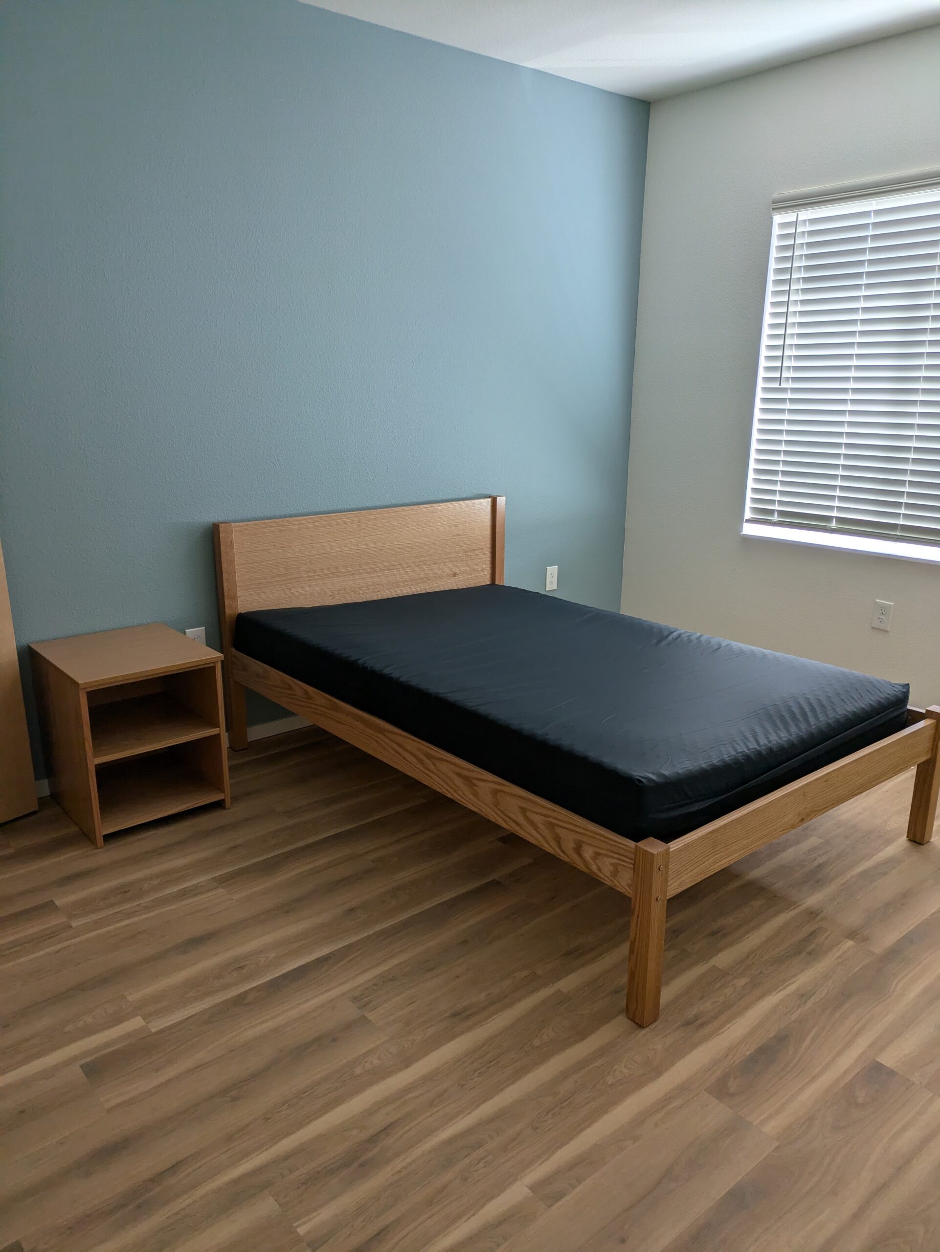 The bedroom of a new Solid Ground apartment, which is furnished with a wooden bed frame with a dark mattress cover and a matching nightstand. The room has light wood-laminate floors and a blue-gray accent wall behind the bed.