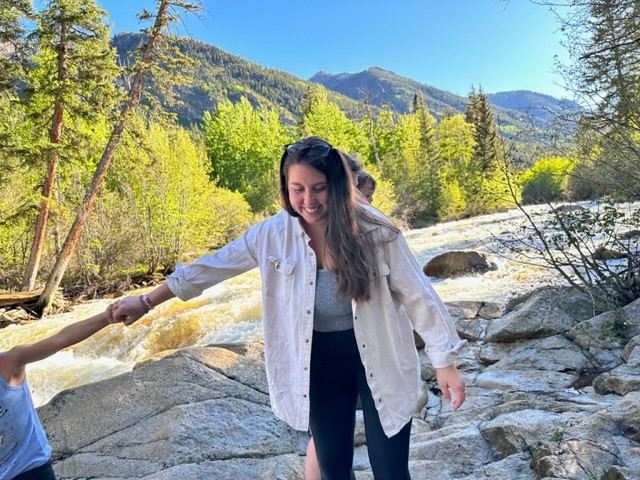 Uma jovem de longos cabelos castanhos, Silvia, sorri enquanto está de pé sobre grandes rochas à beira de um rio caudaloso, segurando a mão de uma criança para guiá-la. A cena é uma paisagem montanhosa ensolarada com álamos e pinheiros ao fundo.