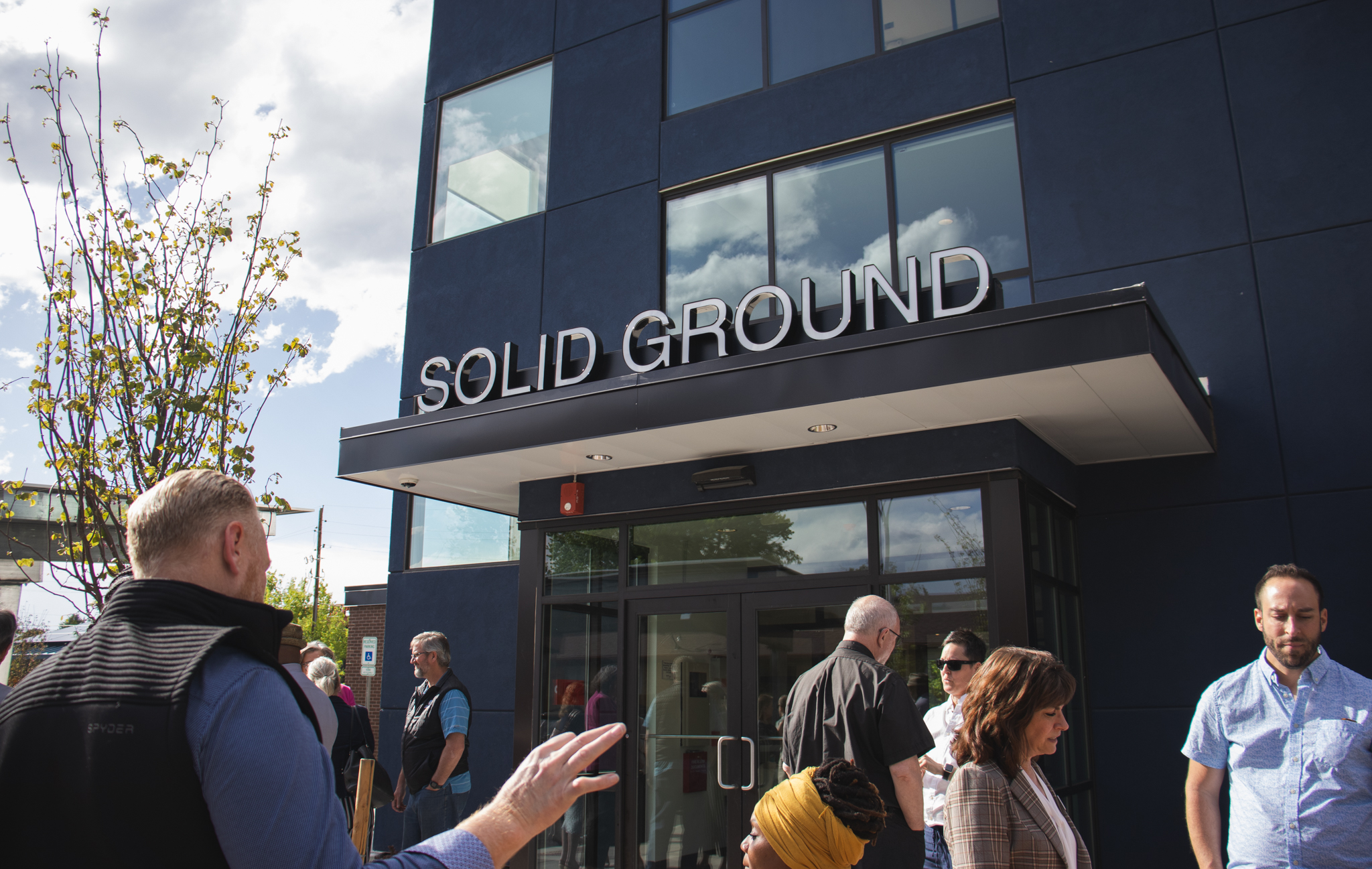 A crowd of people gathers outside the main entrance of the modern, dark-blue "Solid Ground" building. A man in the foreground gestures while speaking to the group, who are mingling before an event.