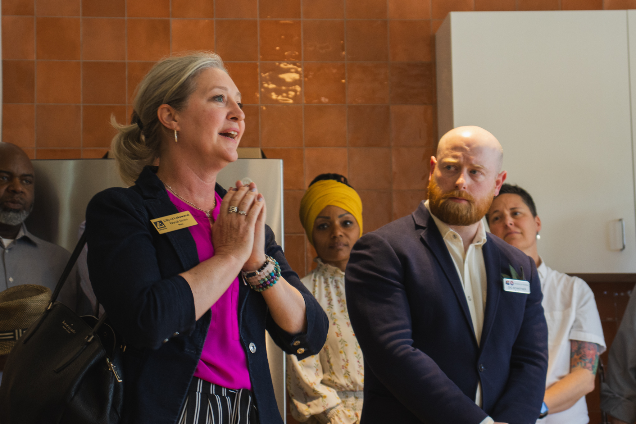 A woman with blonde hair, wearing a black blazer and a bright pink shirt, speaks passionately with her hands clasped. A diverse group of people, including a bald man with a red beard, stand around her, listening intently during the Solid Ground opening event.