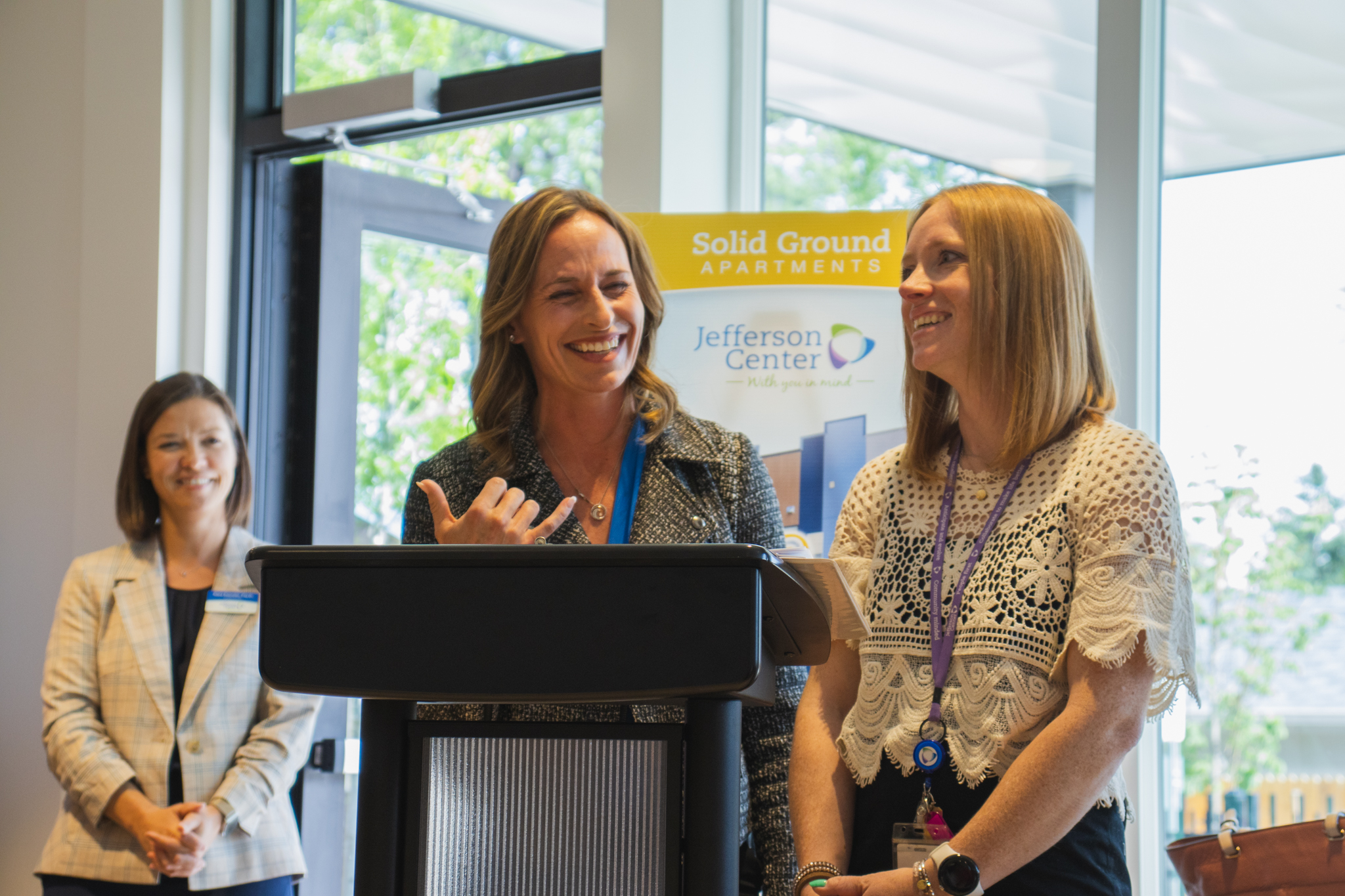 Three women smile while standing at a podium during the opening ceremony for Solid Ground Apartments. The woman at the podium gestures as she speaks, while her colleagues look on. A banner for the apartments and Jefferson Center is in the background.