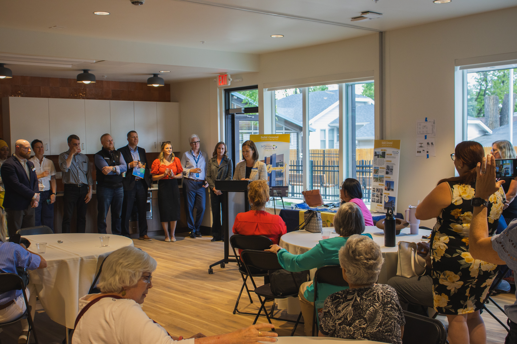 A wide-angle view of the Solid Ground opening ceremony. A speaker stands at a podium addressing a seated audience, while a group of dignitaries stands behind them in front of a bright window.