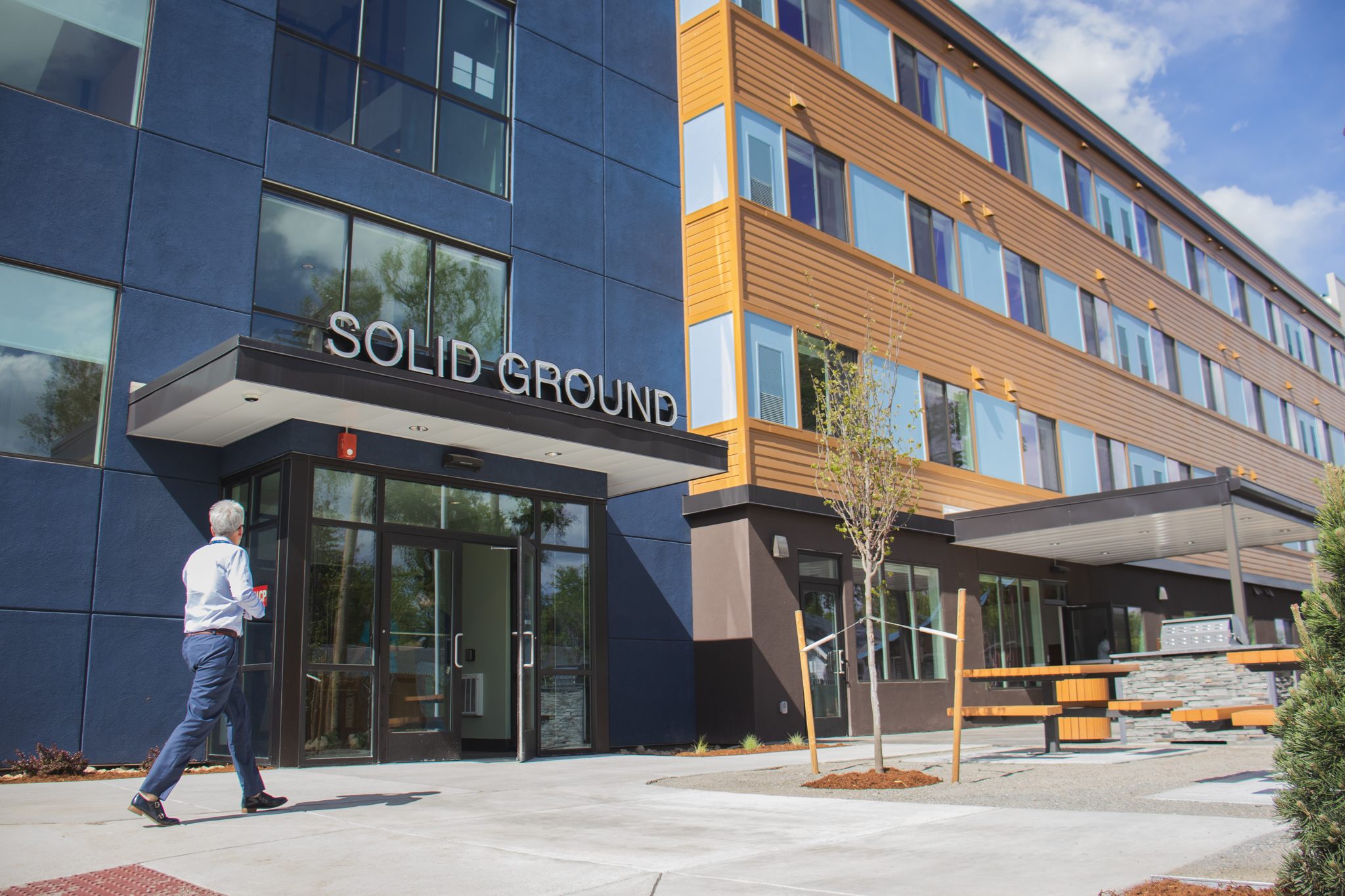 The exterior of the modern Solid Ground apartment building on a sunny day. The multi-story building features a mix of dark blue and wood-paneled siding, with the name "Solid Ground" in large letters over the main entrance.