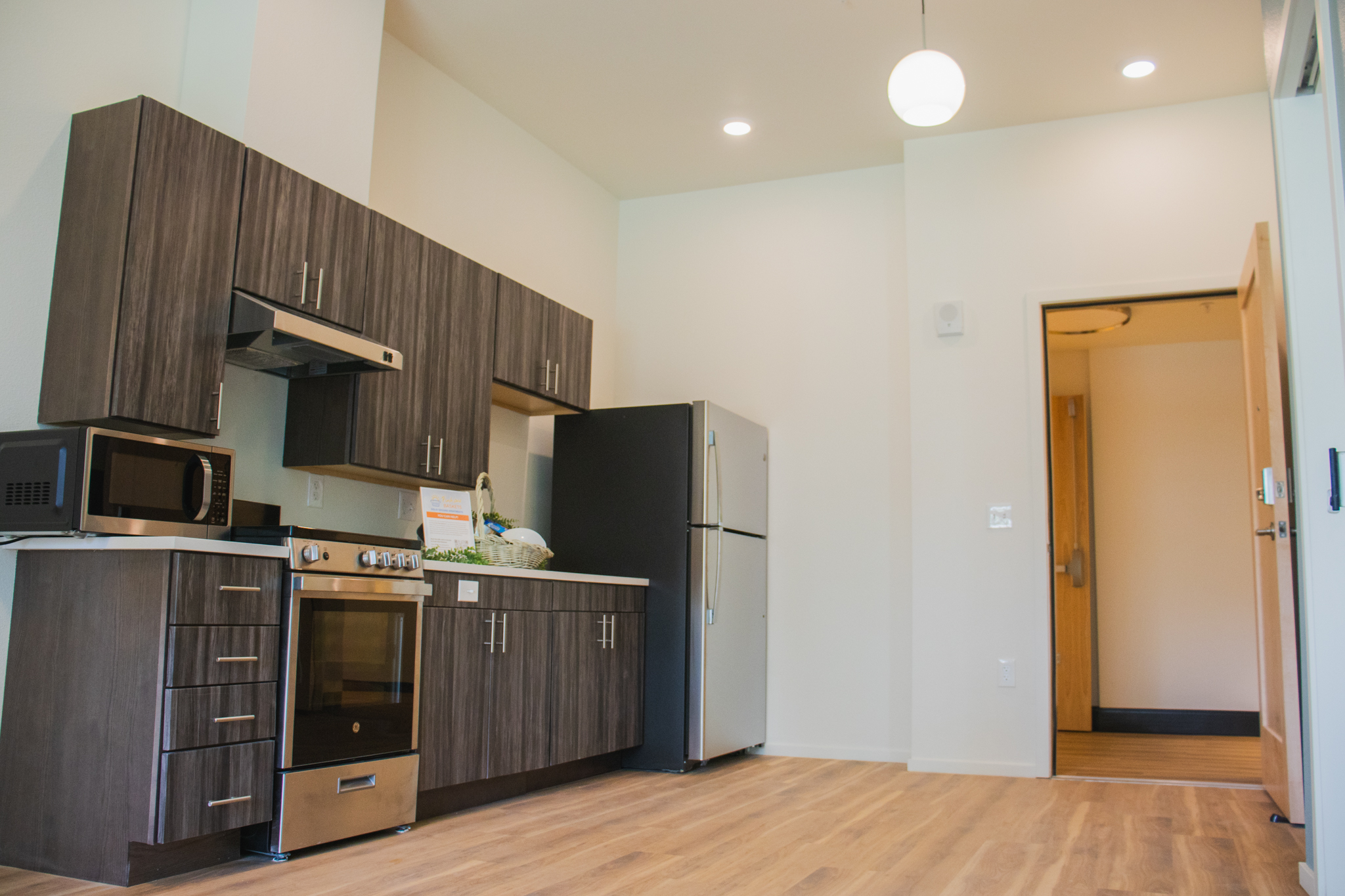 The new kitchen in a Solid Ground apartment, featuring modern, dark wood-grain cabinets, stainless steel appliances, and light wood-laminate flooring. A small welcome basket sits on the counter.