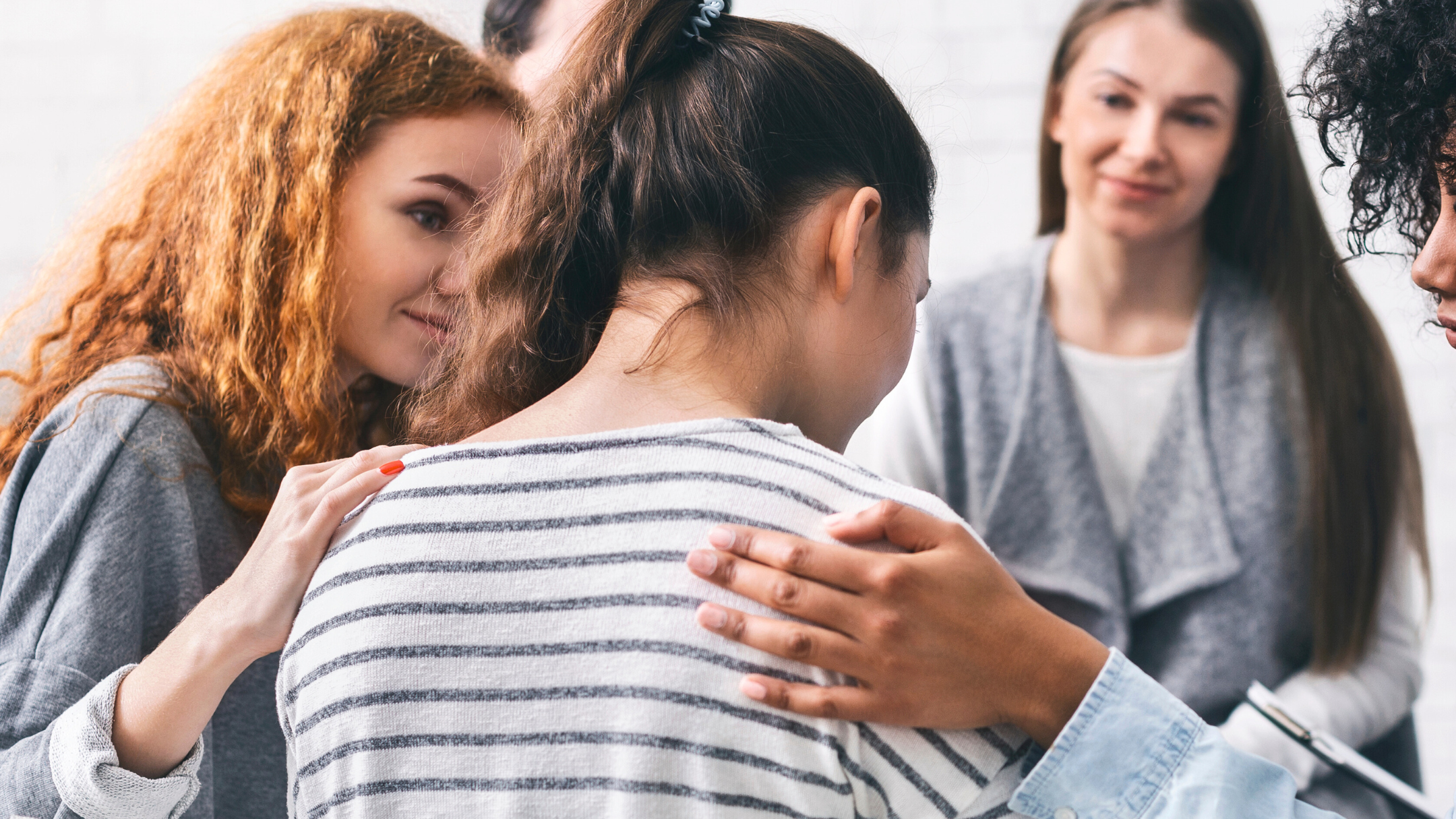 In a support group setting, a person seen from behind is comforted by fellow members who place supportive hands on their shoulders and back. A woman with long red hair leans in with an empathetic expression, creating a powerful image of community and care.