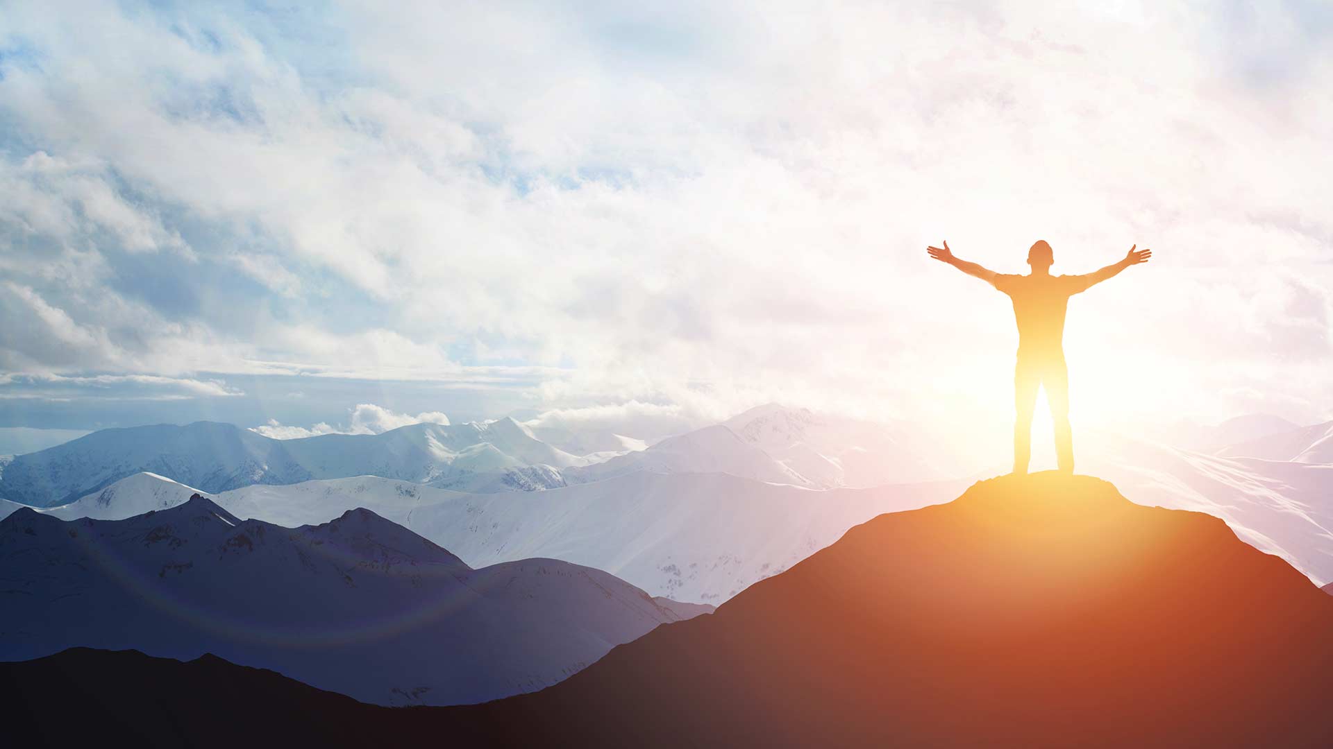 Силуэт человека стоит на вершине горы на рассвете, раскинув руки в триумфальной позе. Вдали виднеется обширный заснеженный горный хребет.