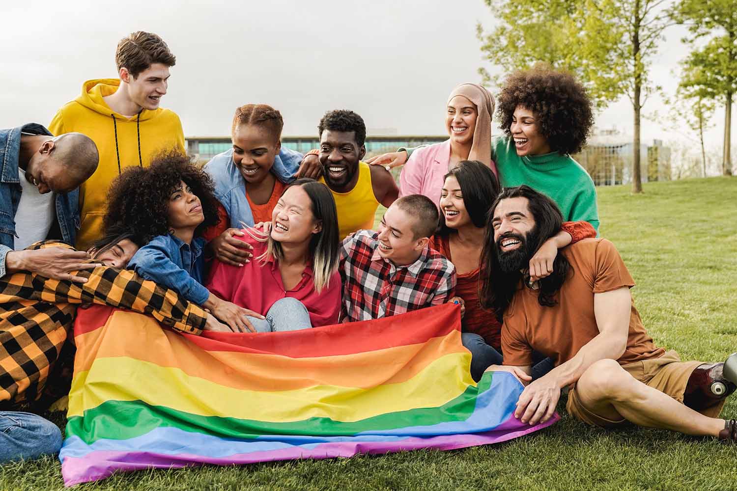 Un grupo numeroso y diverso de jóvenes amigos ríe y sonríe sentados en el césped de un parque. Una bandera arcoíris del Orgullo se extiende frente a ellos, y el grupo incluye personas de diversas razas y capacidades.