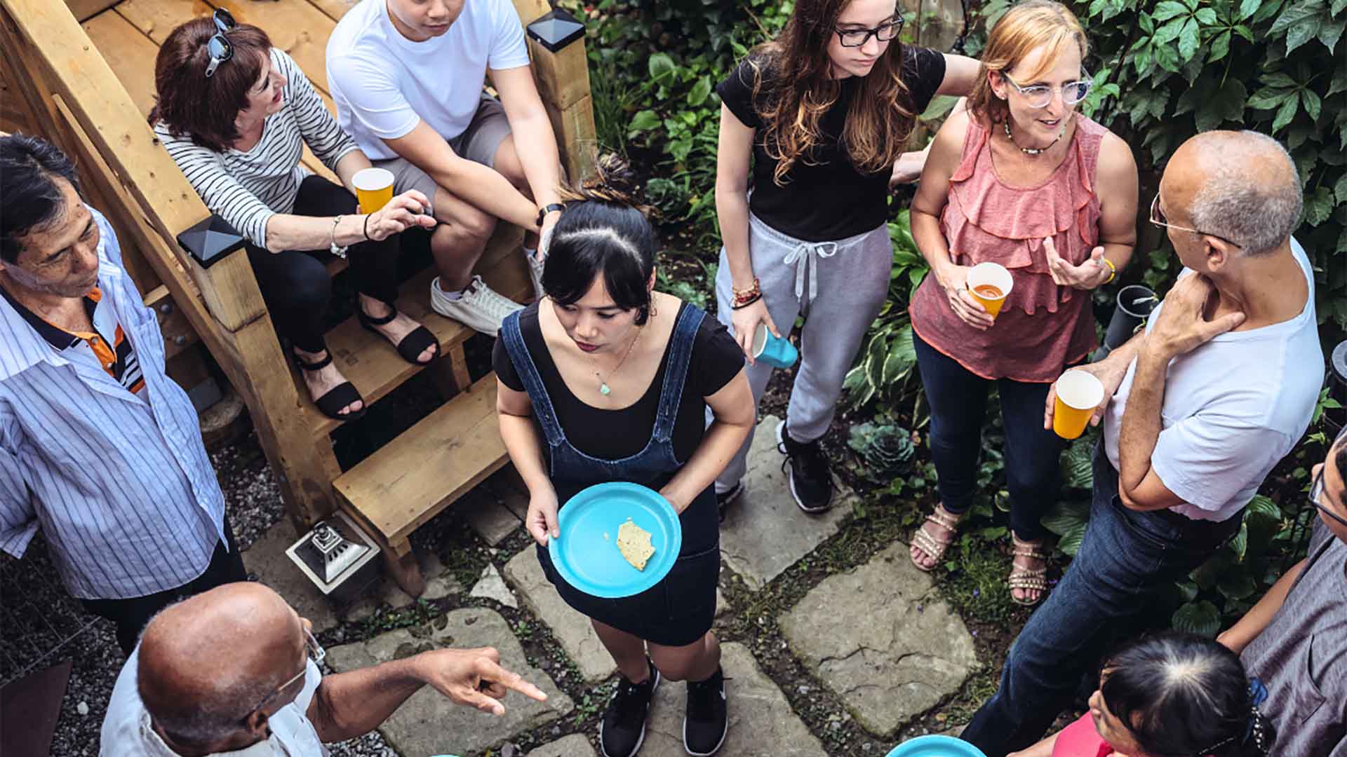 Vu d'en haut, un groupe diversifié et multigénérationnel est réuni sur une terrasse, discutant et socialisant. Debout ou assis, ils forment un cercle informel, tenant des assiettes et des tasses.