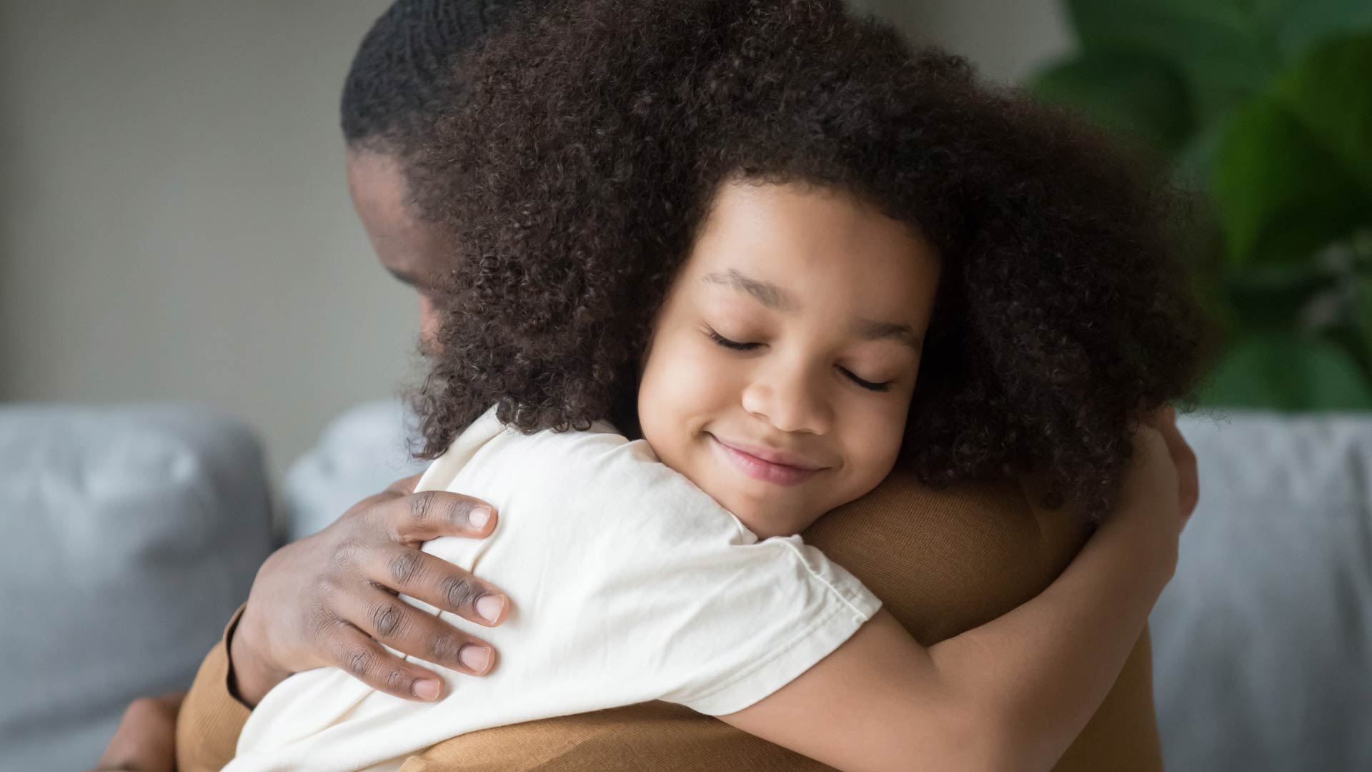 A young child with long, curly hair has a peaceful and contented smile with their eyes closed as they are held in a tight, loving hug by their father.