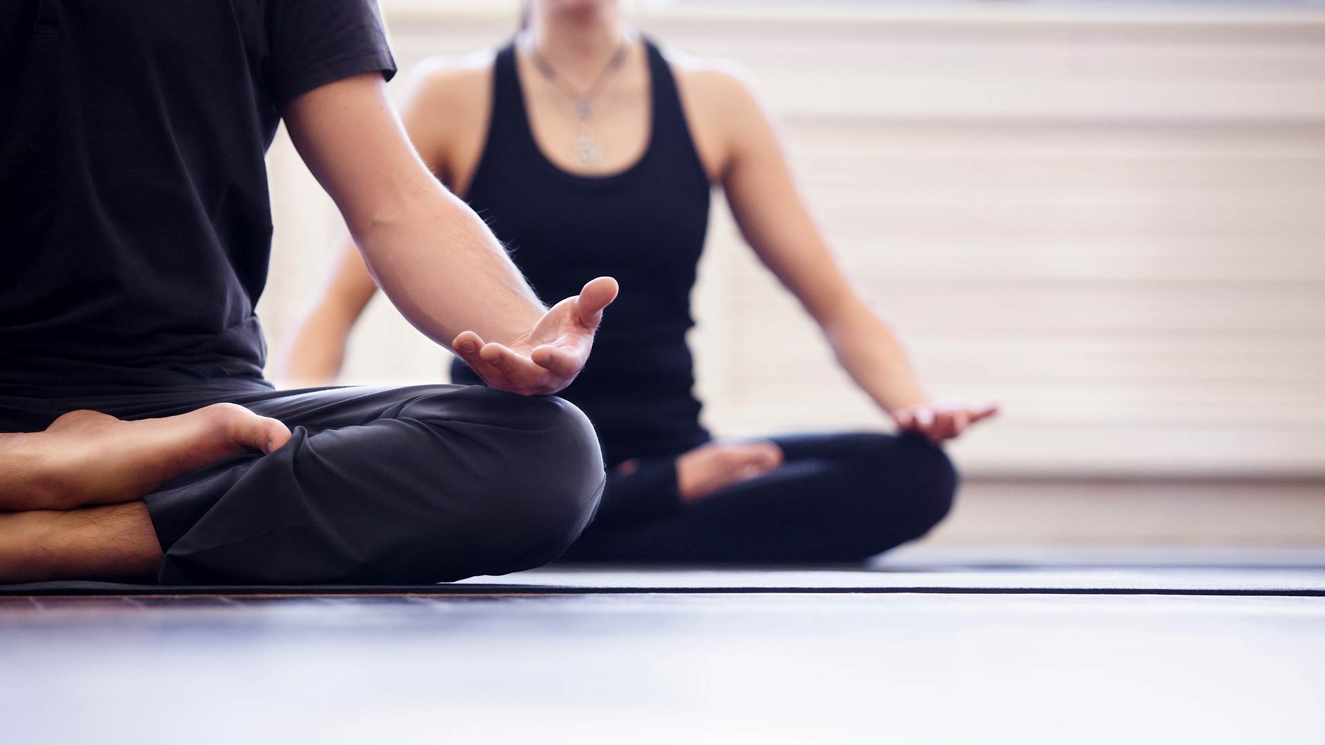 Primer plano, vista desde abajo, de dos personas sentadas en postura meditativa con las piernas cruzadas sobre colchonetas de yoga. El enfoque se centra en la persona en primer plano, cuya mano descansa sobre la rodilla con la palma hacia arriba, creando una escena de paz y consciencia plena.
