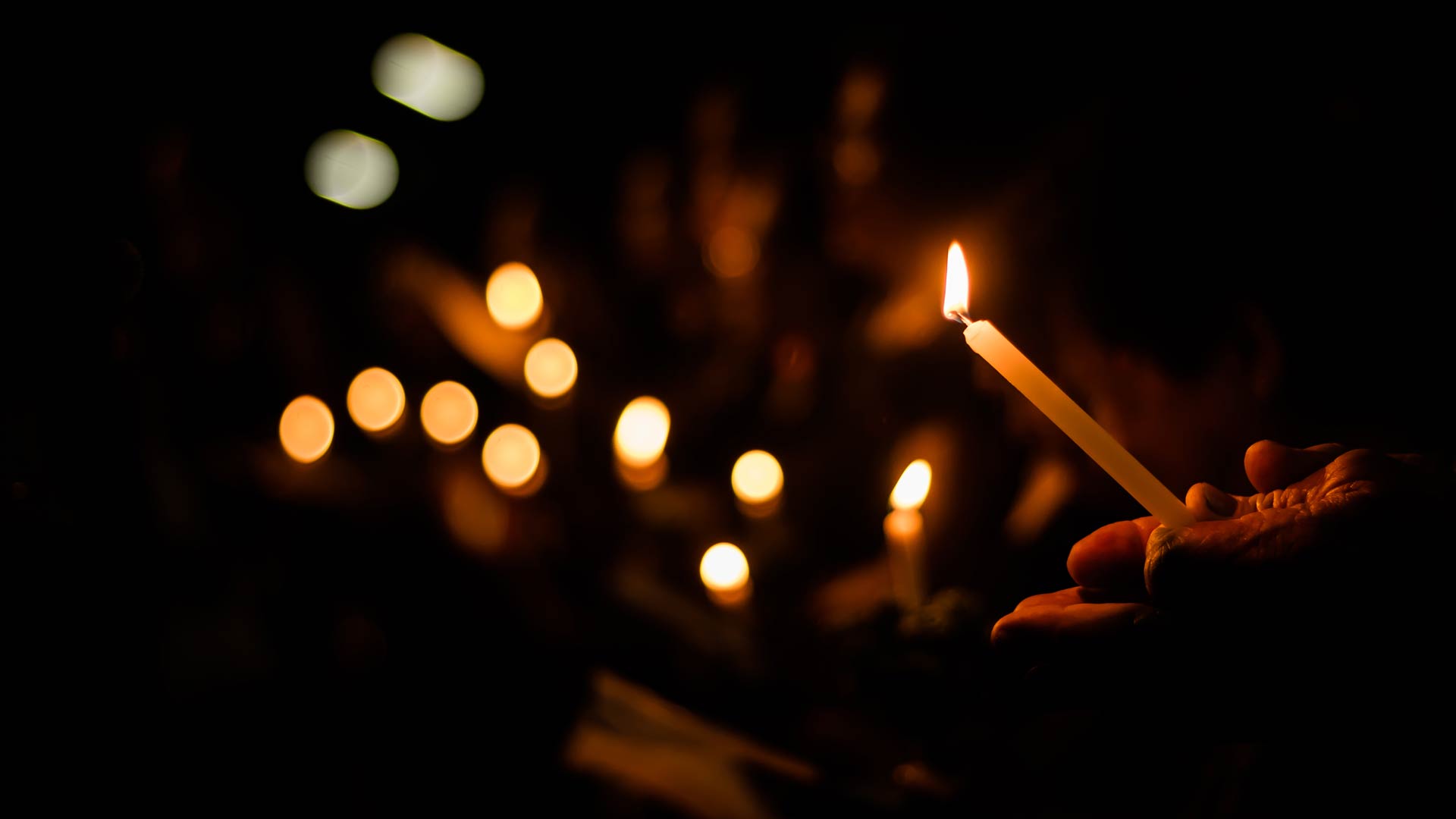 A person's hand holds a thin, lit candle in the dark during a vigil. The single flame is in focus, while the out-of-focus glows of many other candles are visible in the background.