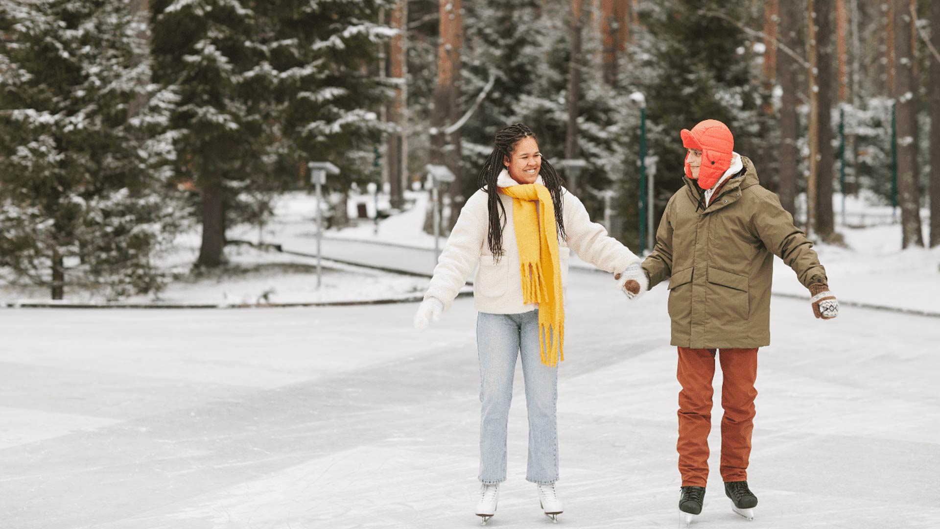 Una joven pareja se toma de la mano mientras patina sobre hielo en una pista al aire libre. La mujer, negra y con una bufanda amarilla brillante, le sonríe. Están rodeados por un paisaje nevado con árboles de hoja perenne.