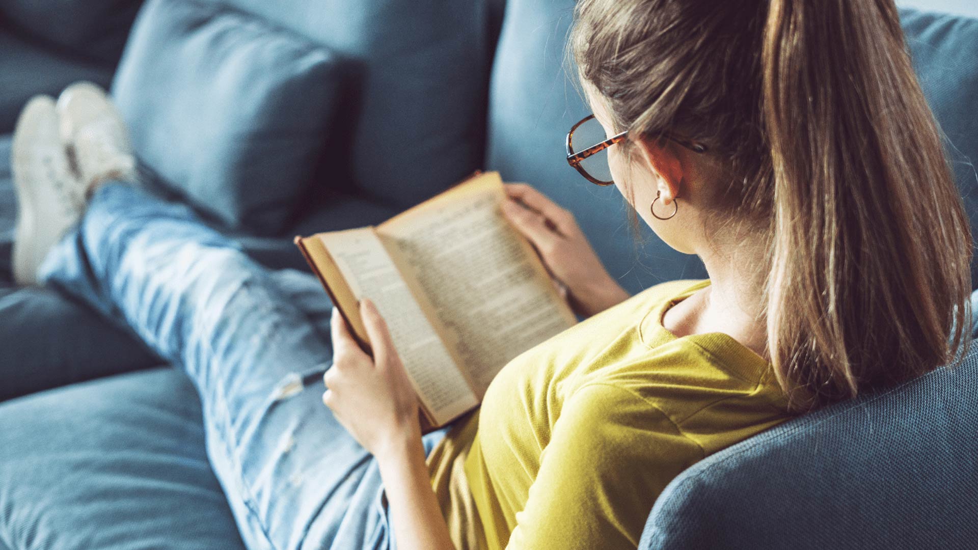 Vista da sopra la sua spalla, una giovane donna con gli occhiali e i capelli raccolti in una coda di cavallo legge un libro mentre è sdraiata su un divano blu. Indossa una maglietta gialla e jeans, e tiene i piedi appoggiati in una posizione rilassata.