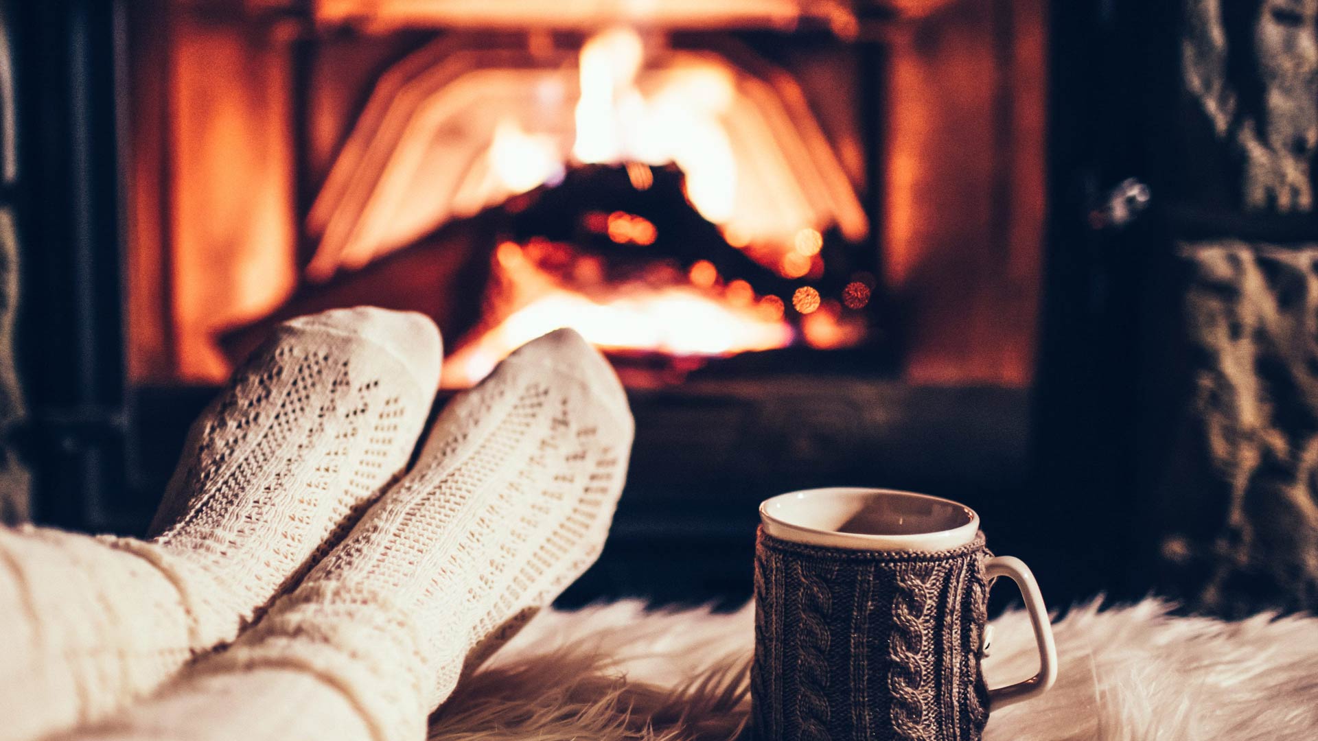 A first-person view of a person's feet in cozy, cream-colored knit socks, propped up on a fuzzy white rug in front of a glowing fireplace. A mug wrapped in a knit cozy sits next to them