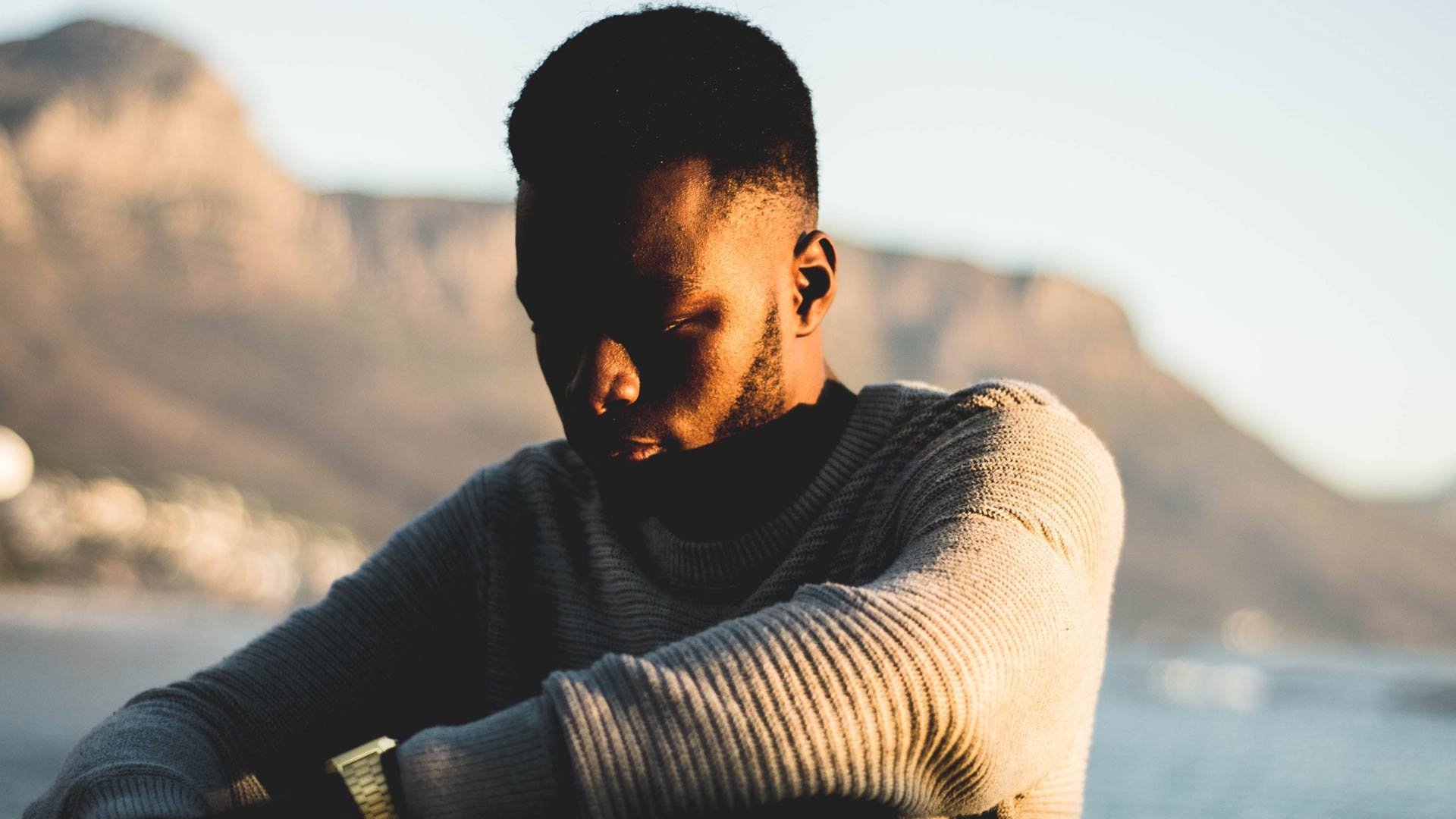 A young Black man with his head bowed in quiet contemplation, lit by the warm light of a setting sun. A mountain is visible in the soft-focus background