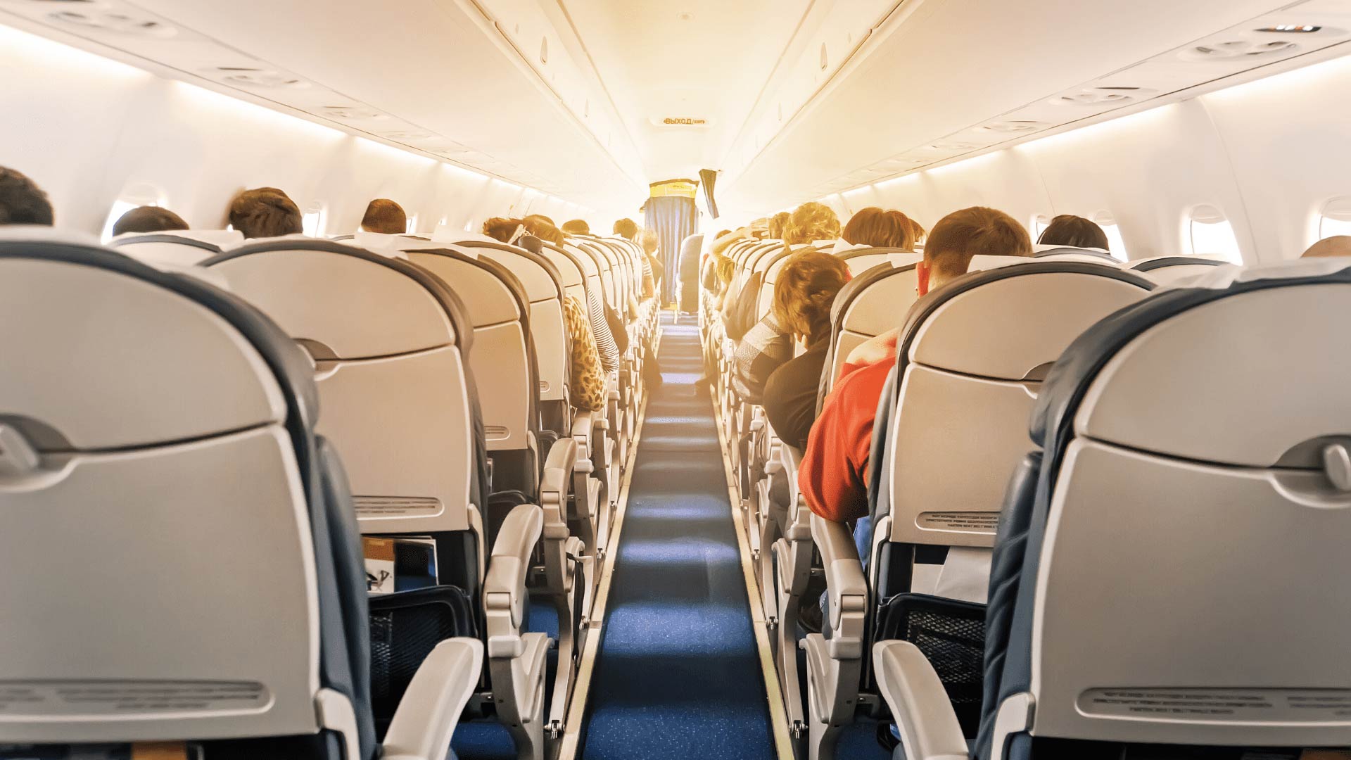 A view looking down the center aisle from the back of a full passenger airplane. Passengers are visible in their seats, and bright sunlight is streaming into the cabin from the front.