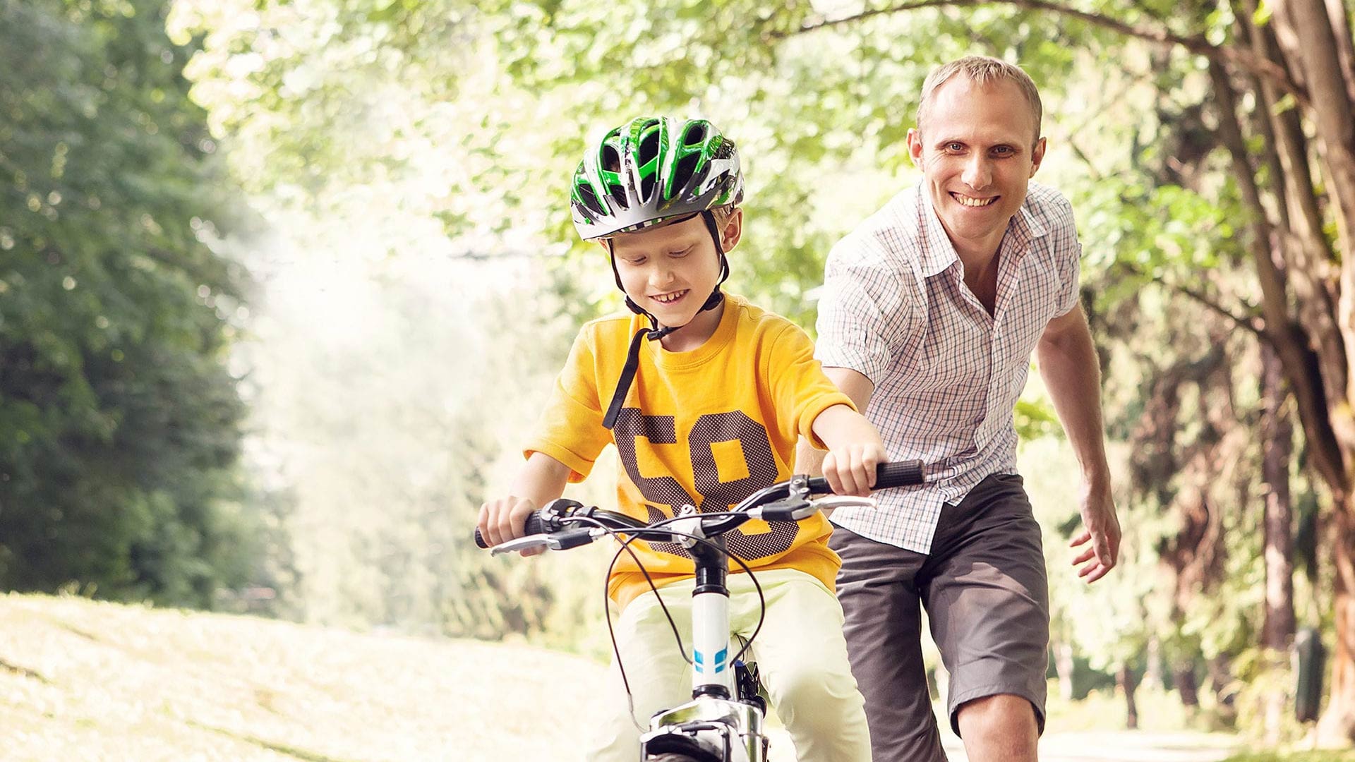 A father smiles proudly as he helps his young son learn to ride a bike on a path through a park. The boy, wearing a yellow shirt and a green helmet, concentrates on steering as his father stays close behind to support him on a sunny summer day.