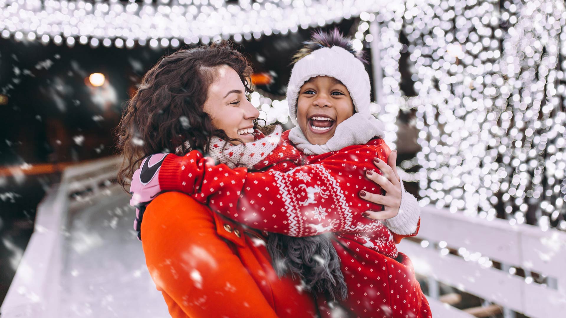 A woman with curly dark hair warmly embraces a young, laughing child in a festive, snowy, nighttime scene. The child, wearing a red patterned sweater and a white pom-pom hat, has a look of pure joy. Snowflakes fall around them as they stand in front of a glowing wall of out-of-focus holiday lights.