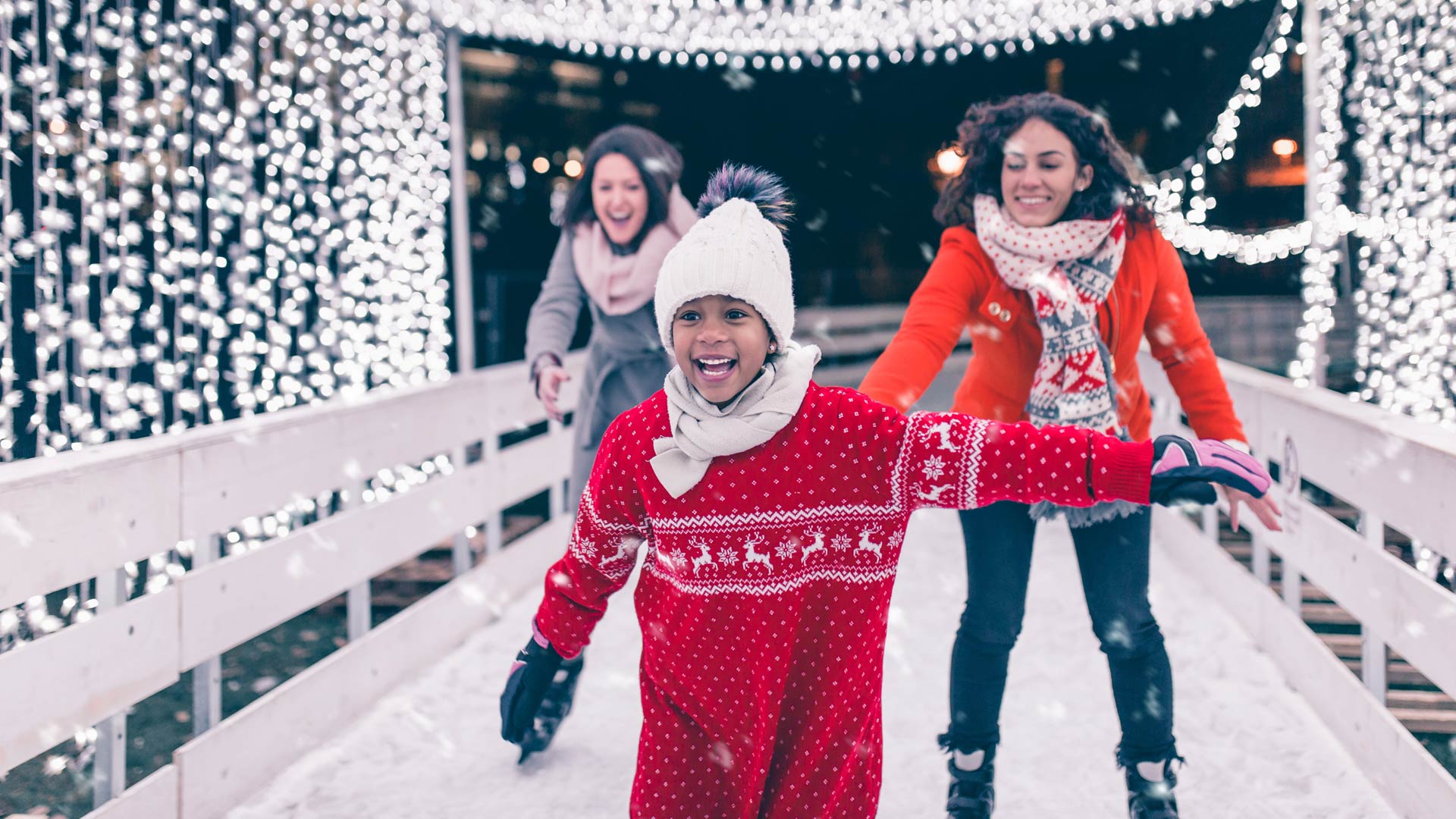 Un jeune enfant, vêtu d'un pull rouge et d'un bonnet blanc, sourit joyeusement en patinant sur une patinoire extérieure. Deux femmes souriantes patinent derrière, profitant toutes d'une soirée festive sous les illuminations de Noël.