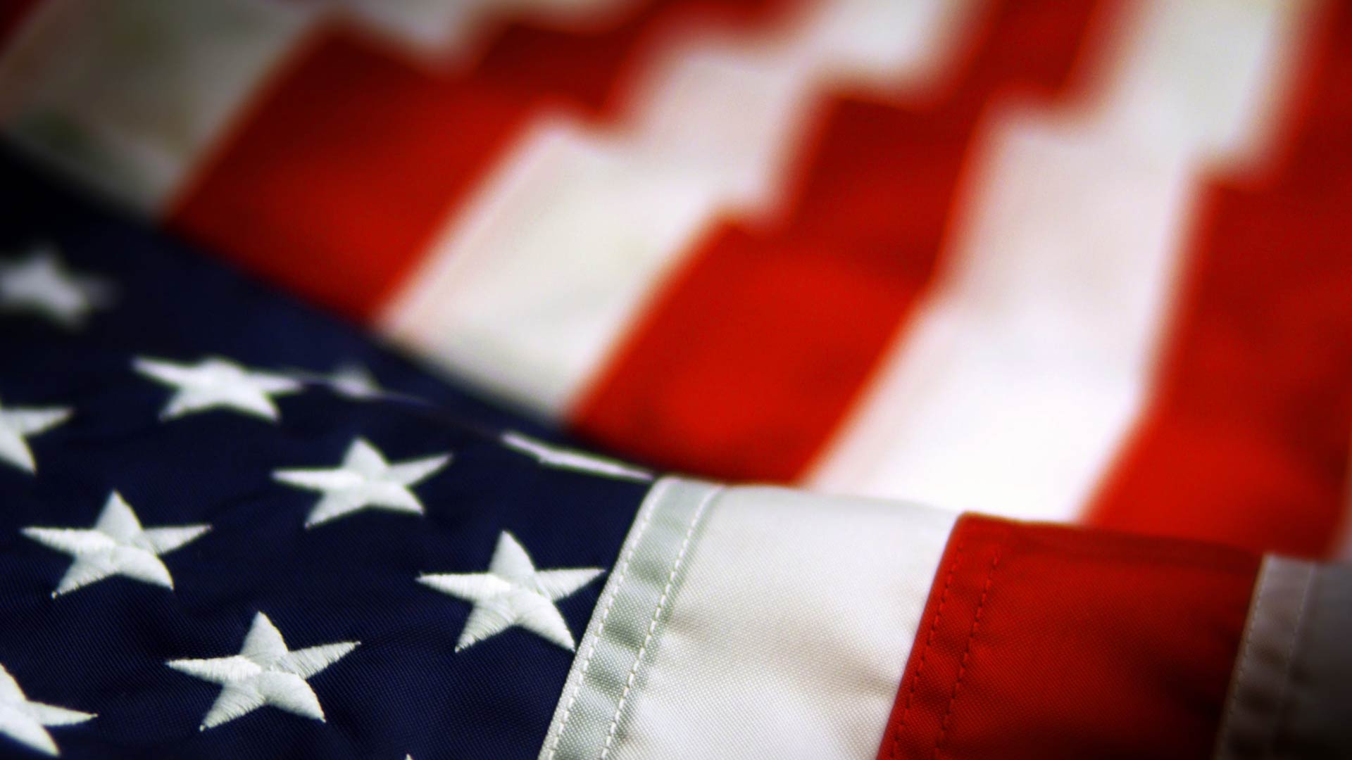 An artistic close-up of the American flag. The blue field and white stars are in sharp focus in the foreground, while the red and white stripes are softly blurred as they wave in the background.