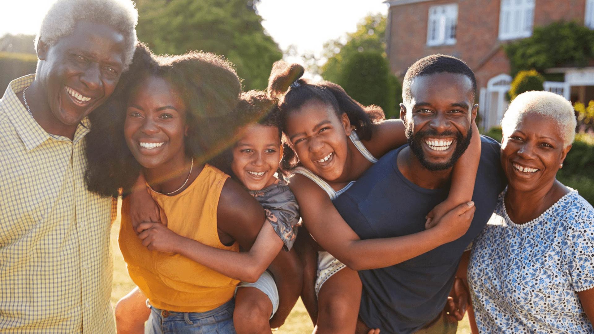 A multi-generational Black family, including grandparents, parents, and two children, smile and laugh together outdoors on a sunny day. The mother and father give their two laughing daughters piggyback rides, creating a joyful and loving family portrait.