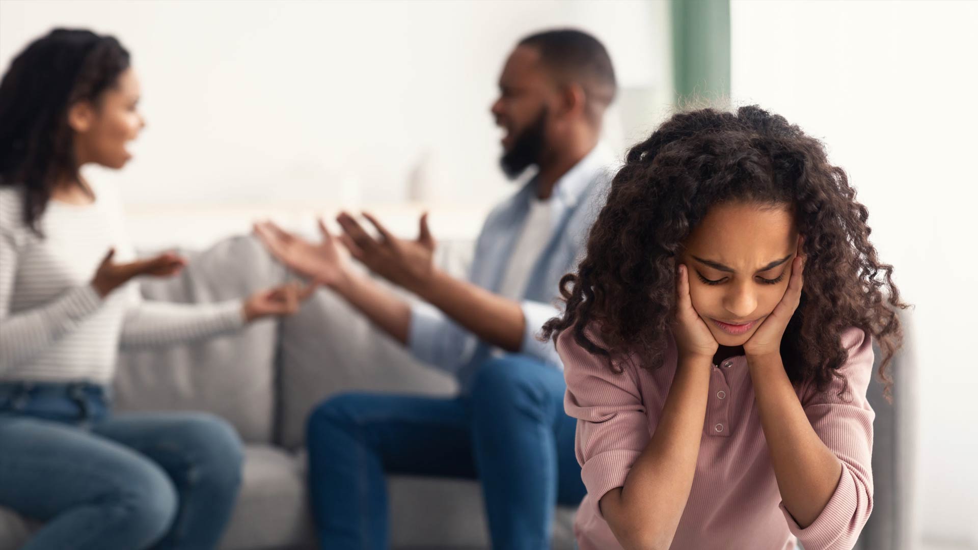 A young Black girl with a distressed expression holds her head in her hands, while in the blurred background, her parents sit on a couch and argue.