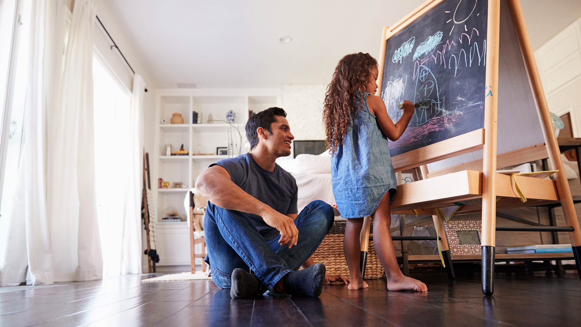 Uma jovem de cabelos longos e cacheados desenha em um cavalete de quadro-negro em uma sala de estar ensolarada. Seu pai está sentado no chão ao lado dela, observando com um sorriso caloroso e orgulhoso.