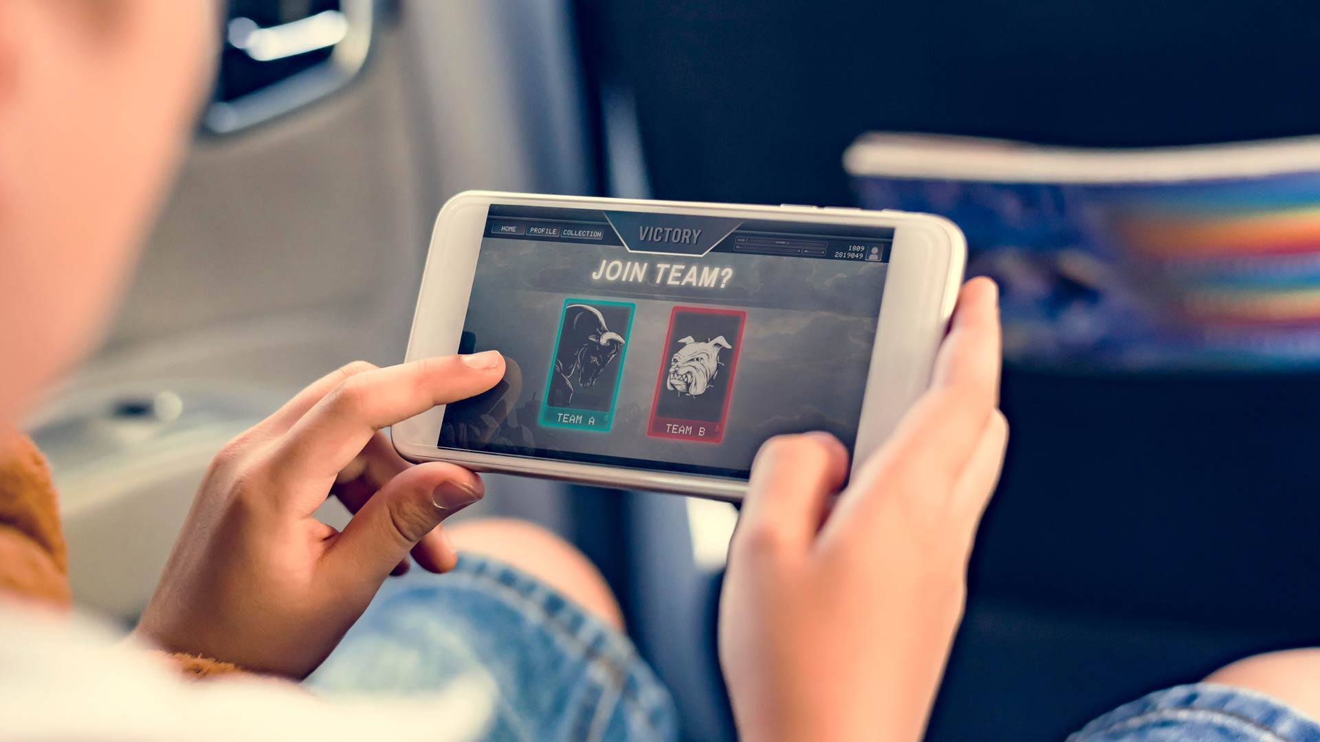 A close-up, over-the-shoulder view of a child sitting in a car while playing a game on a smartphone. The screen shows a "Victory" message and prompts the player to "Join Team?" with two different team logos to choose from.