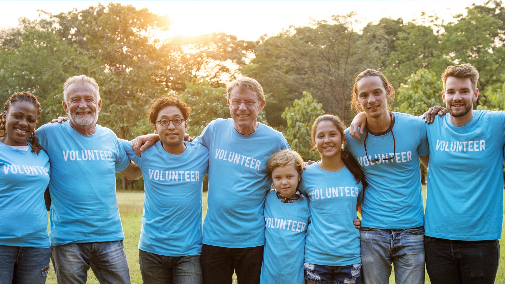 A diverse group of smiling volunteers, ranging in age from a young child to seniors, stand with their arms around each other in a park at sunset, all wearing light blue t-shirts with the word "VOLUNTEER"