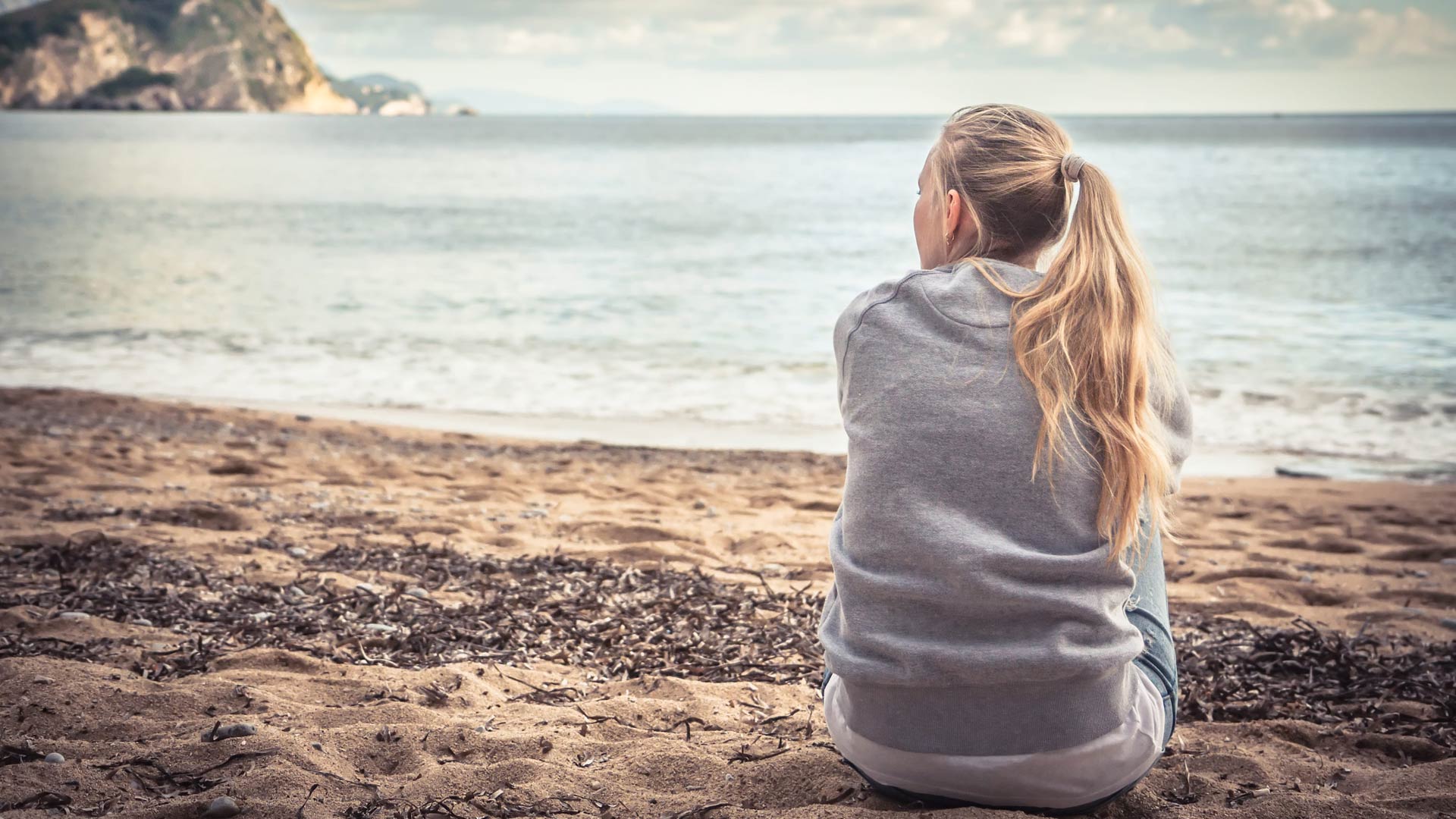 A young woman with blonde hair in a ponytail is seen from behind, sitting alone on a sandy beach. She wears a gray sweatshirt and looks out at the ocean and a distant, hilly shore under a cloudy sky.