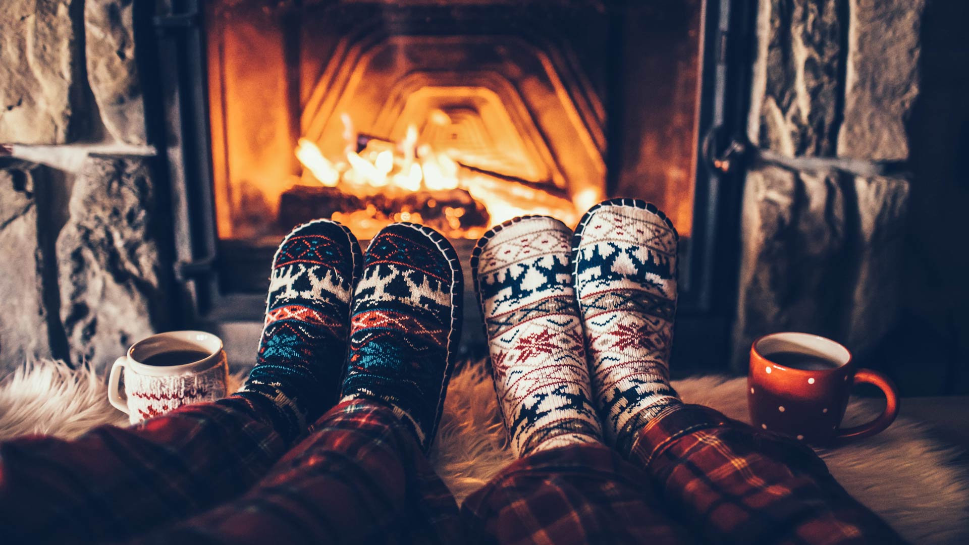 A first-person view of two people's feet, clad in patterned winter socks and red plaid pajamas, propped up in front of a roaring fireplace. They are relaxing on a soft rug with mugs of a hot beverage