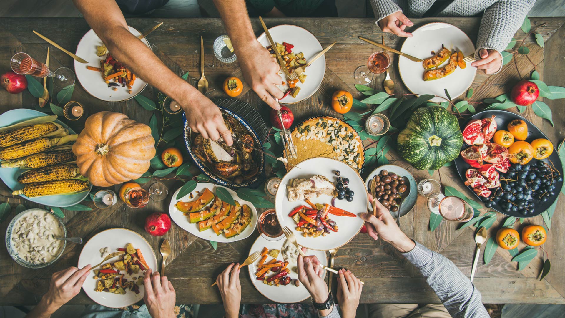 An overhead view of a rustic wooden dinner table laden with a Thanksgiving feast. Multiple hands are reaching to serve themselves from platters of roasted turkey, squash, corn on the cob, and pie. The table is decorated with pumpkins, pomegranates, and greenery, creating a scene of communal celebration.