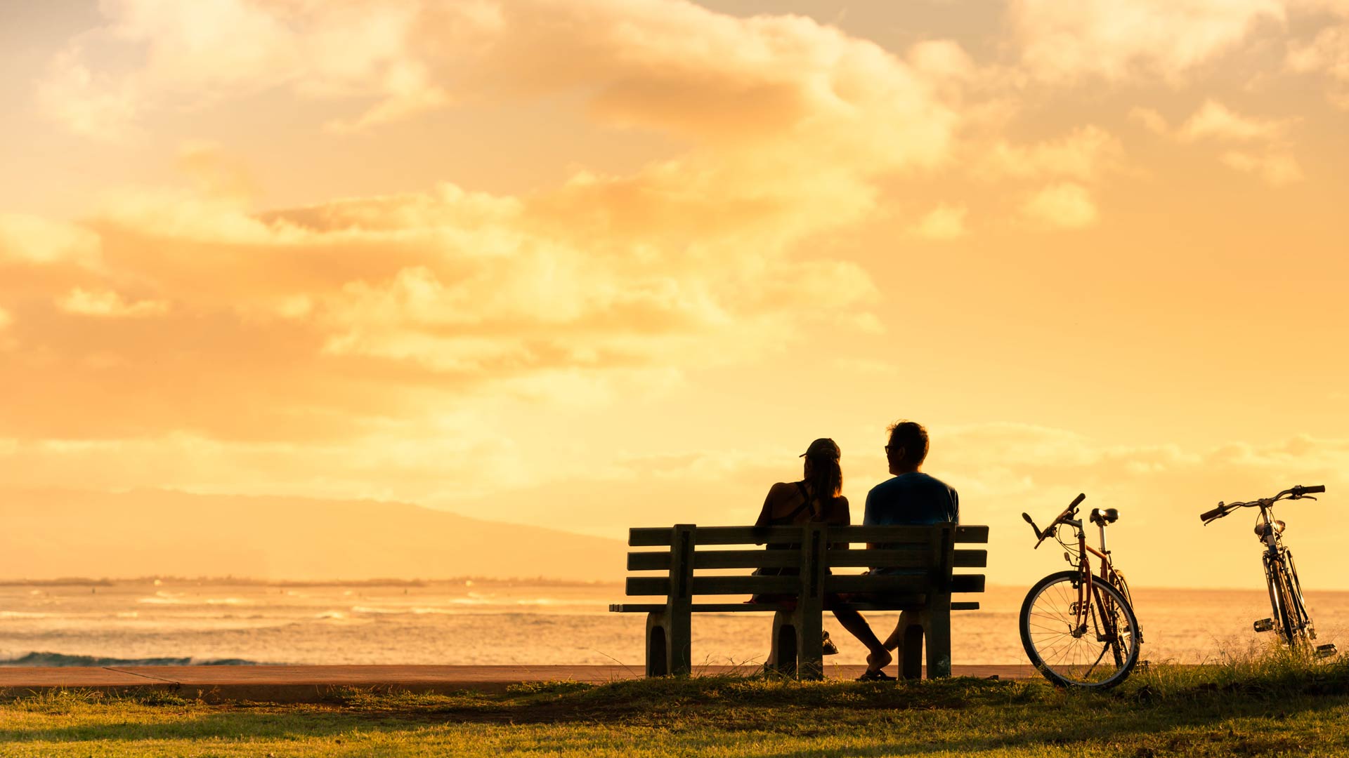 Un couple se dessine sur un banc de parc, contemplant l'océan sous un coucher de soleil chaud et doré. Deux vélos sont garés dans l'herbe à côté d'eux.