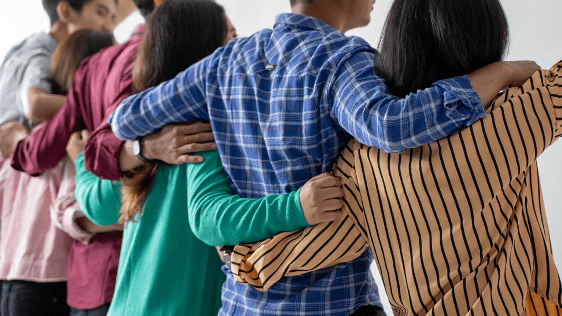 Eine gemischte Gruppe von Menschen steht in einer Reihe (von hinten betrachtet) und hat ihre Arme um die Schultern der anderen gelegt, sodass eine durchgehende Kette der Unterstützung entsteht.