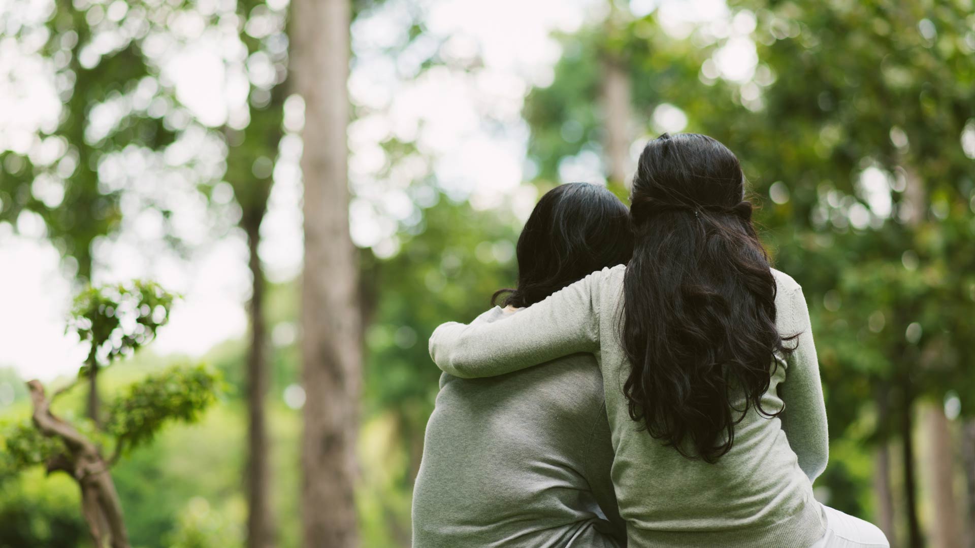 Seen from behind, two people with long dark hair sit closely together in a park. One person has their arm wrapped around the other's shoulders in a gesture of comfort and support.