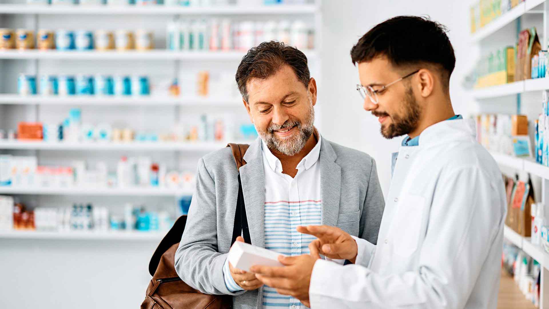A friendly male pharmacist in a white coat explains a medication to a smiling, middle-aged male customer. The pharmacist holds a small white box and points to it as the customer looks on attentively.