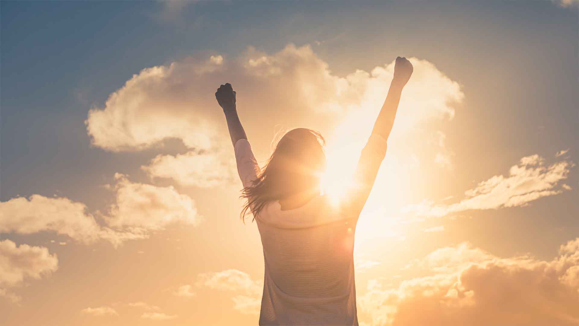 Viewed from behind, a woman stands with her arms raised in a triumphant, victorious pose. She is silhouetted against a bright, golden sun and a sky filled with beautiful clouds.