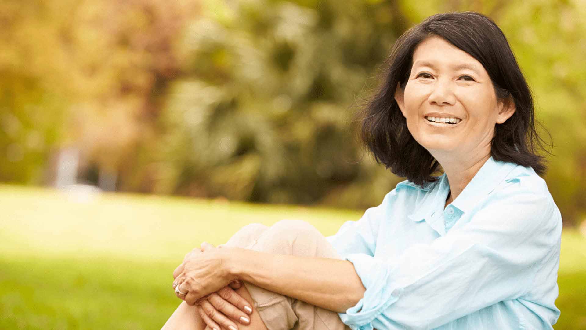 Счастливая женщина среднего возраста из Восточной Азии с короткими темными волосами сидит на травянистой лужайке в парке. Она тепло улыбается, обнимая колени, выглядя расслабленной и довольной.