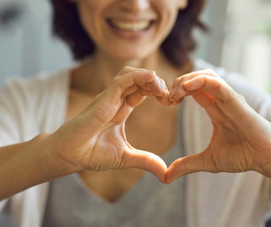 Um close nas mãos de uma mulher enquanto ela as molda em um coração. No fundo de foco suave, seu sorriso caloroso é visível.