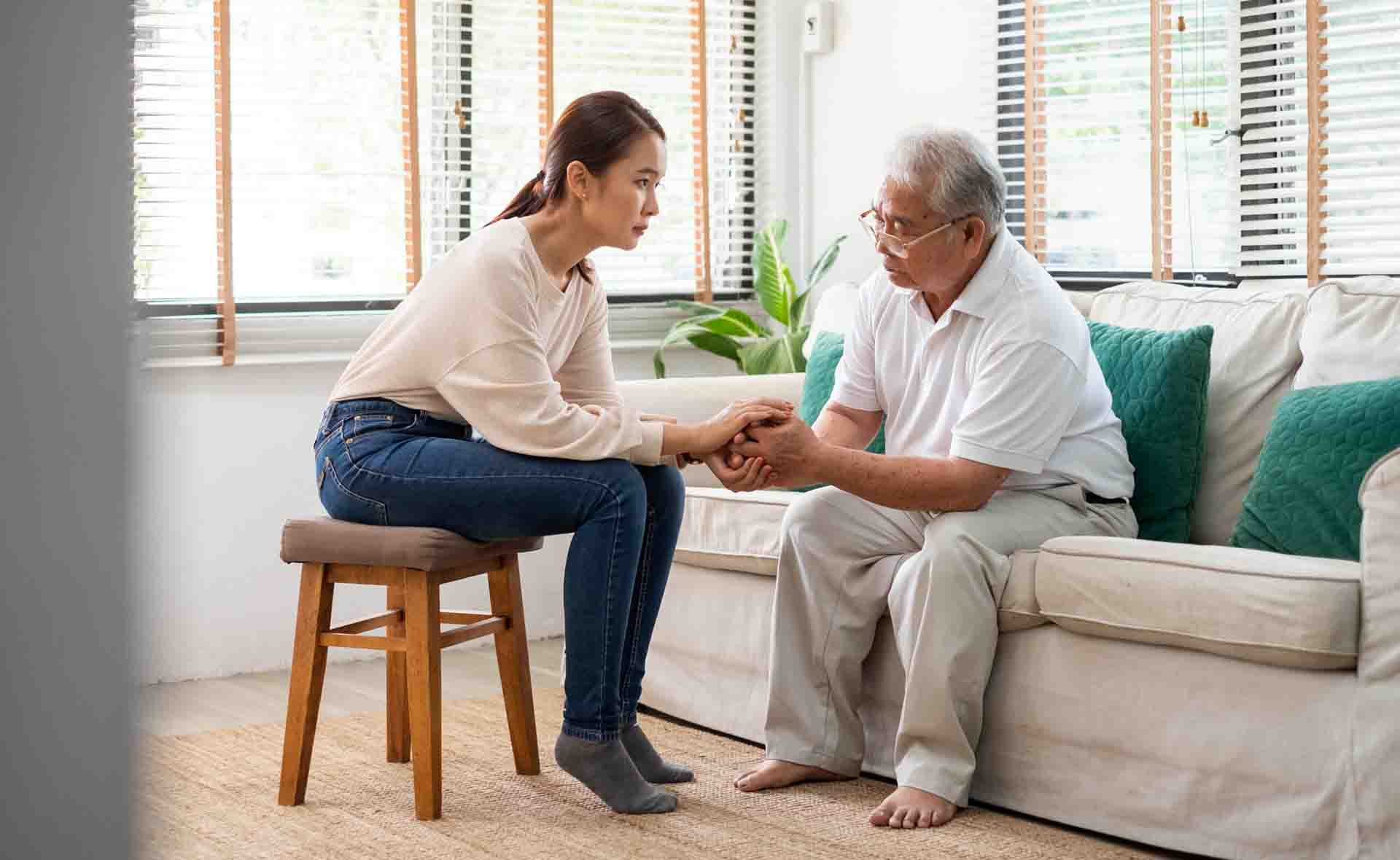 明亮的客厅里，一位年轻女子坐在凳子上，握着一位坐在沙发上的老人的手。他们互相注视着，进行着一场严肃而互相支持的对话。