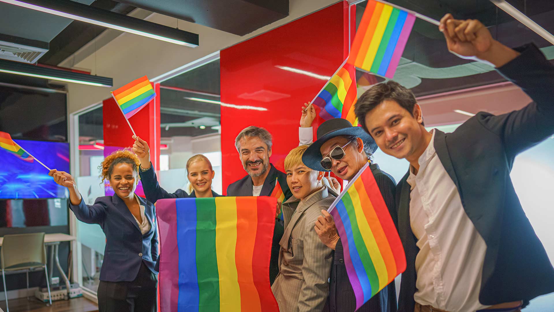 在现代化的办公室里，一群来自不同背景的同事欢声笑语，欢呼庆祝骄傲节。他们手持大大小小的彩虹旗，欢欣鼓舞地表达着对骄傲节的支持与团结。