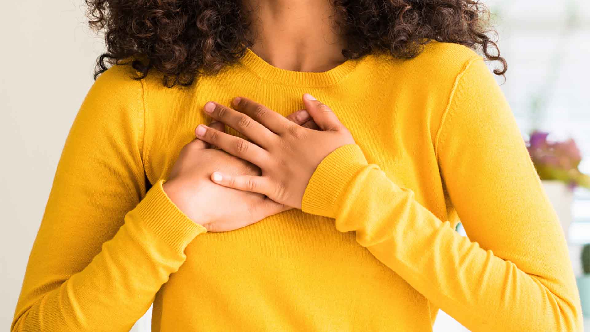 Крупный план женщины в ярко-желтом свитере, которая нежно прижимает обе руки к сердцу в жесте благодарности.